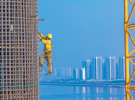 奮戰(zhàn)在百米索塔之上的建設(shè)者身影，與遠(yuǎn)處的高樓大廈遙呼相應(yīng)。